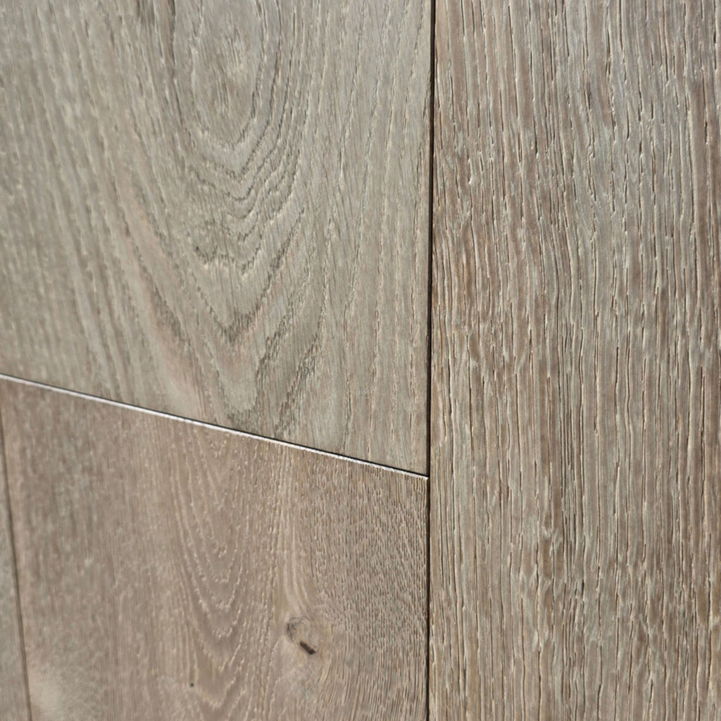 Light oak wood flooring planks with visible grain patterns and natural knots in a warm grayish-brown tone. The flooring in this image is Engineered European Oak Wide Plank Flooring Ember Valley. shown very close up 