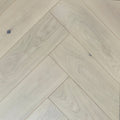 Light natural wood flooring planks arranged in a herringbone pattern, showing pale beige tones and subtle wood grain texture. Engineered European Oak Whistler Ascent - Herringbone