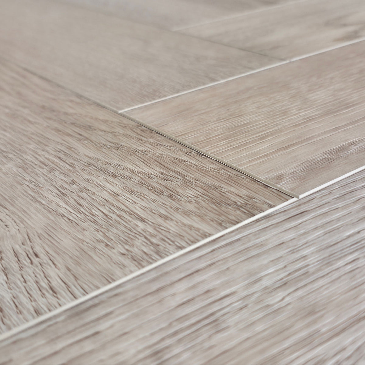Close-up of light oak look SPC flooring planks arranged in a herringbone pattern, showing natural wood grain texture and thin grout lines. Close up of Geneva Mist - Herringbone