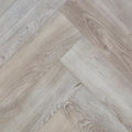 Light gray wood-look flooring planks arranged in a herringbone pattern, showing natural grain and subtle tonal variation. The image is of Highland Ore - Herringbone
