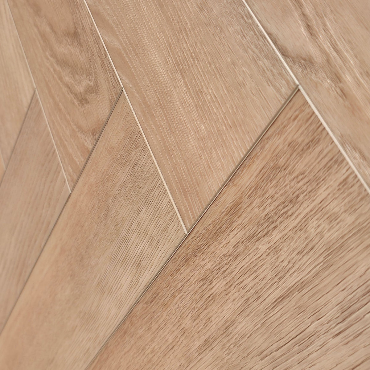 Close-up of light oak wood look SPC flooring planks arranged in a herringbone pattern, showing natural wood grain texture and subtle grout. The flooring shown is Valence Harvest - Herringbone