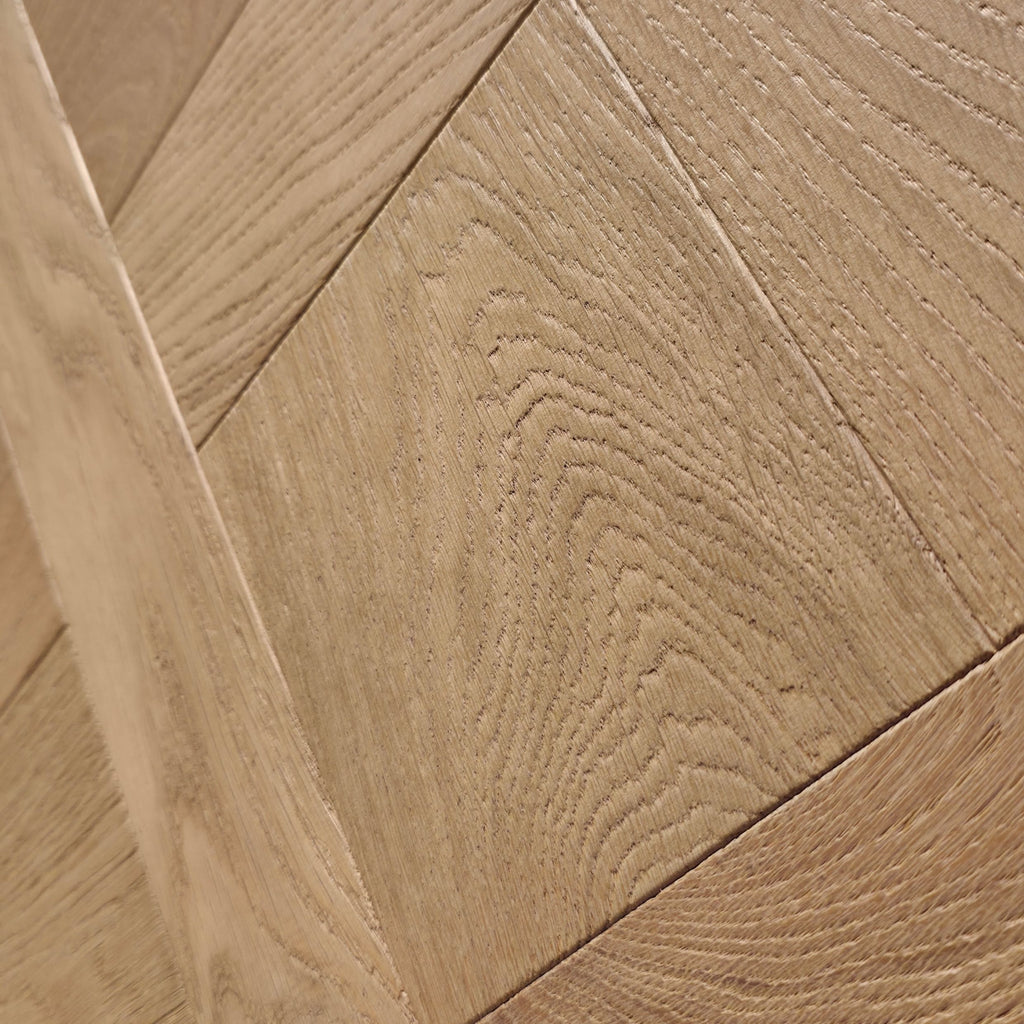 Close-up of light oak wood flooring planks arranged in a herringbone pattern, showing detailed wood grain texture. Engineered European oak Gstaad Glow Parquet Tile is pictured. 