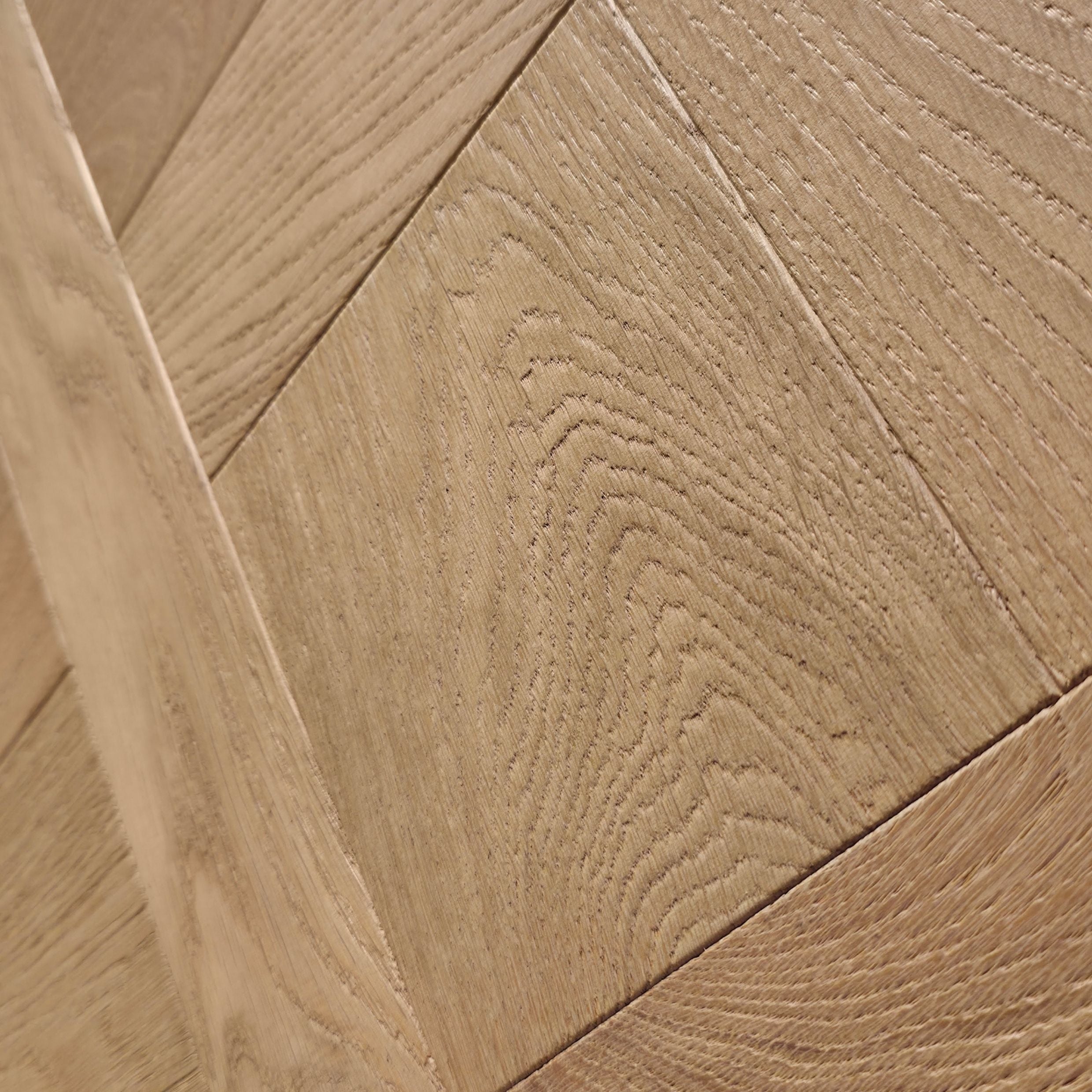 Close-up of light oak wood flooring planks arranged in a herringbone pattern, showing detailed wood grain texture. Engineered European oak Gstaad Glow Parquet Tile is pictured. 