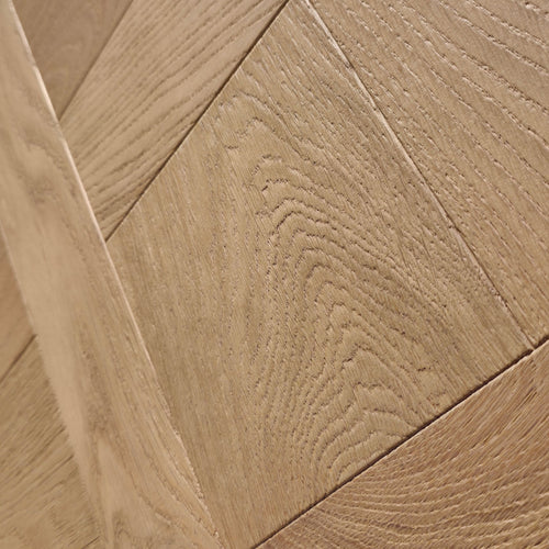 Close-up of light oak wood flooring planks arranged in a herringbone pattern, showing detailed wood grain texture. Engineered European oak Gstaad Glow Parquet Tile is pictured. 